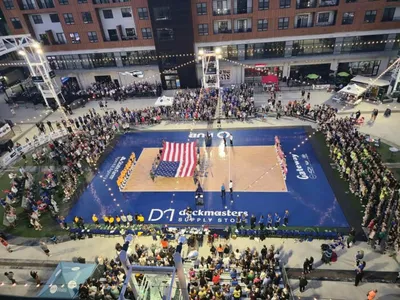 Aerial view of an outdoor volleyball court surrounded by a large crowd, with an American flag displayed at center