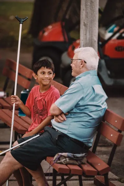 A smiling boy in a red t-shirt holds a pole while chatting with an older man on a wooden bench