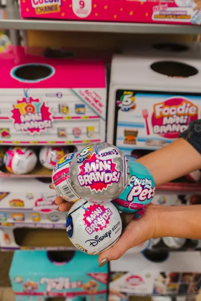 Hand holding three Mini Brands collectible toy balls in front of store display shelves