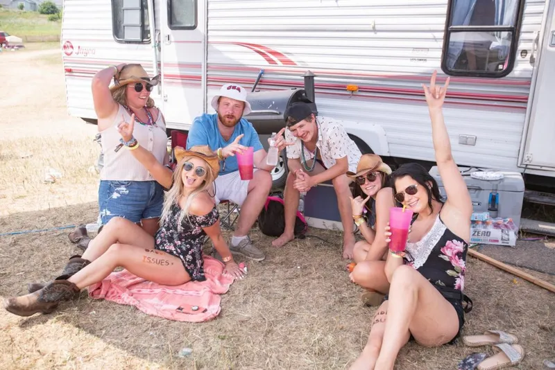 Group of six friends in cowboy hats posing with pink drinks beside a vintage camper trailer