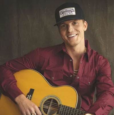 Smiling man in red shirt and trucker hat holding an acoustic guitar