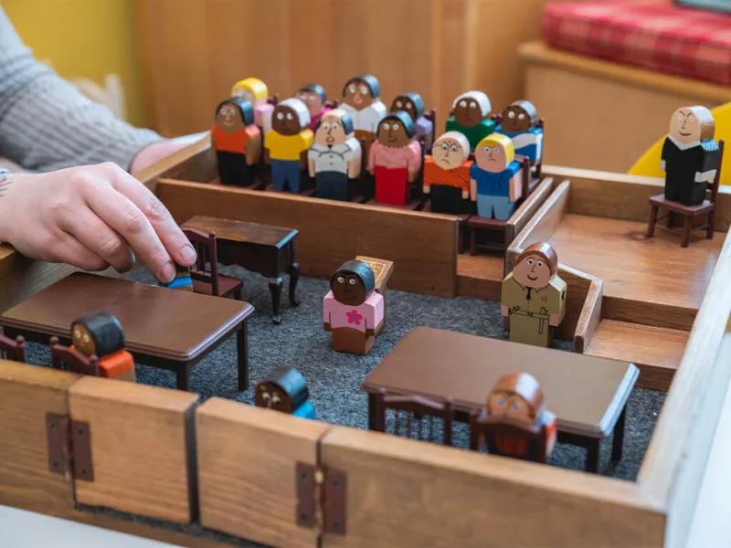 Hand arranging painted wooden figures in a miniature courtroom model with jury box and judge