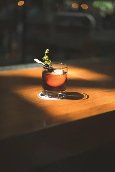 Dark cocktail in a rocks glass garnished with a berry and fresh herb sprig, lit by warm spotlight on a wood bar