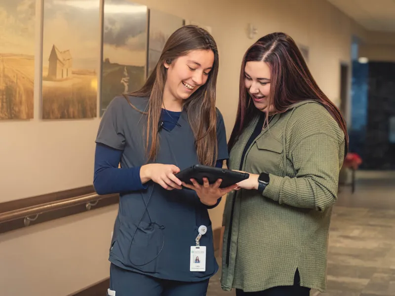 A smiling nurse in scrubs with a badge shows a tablet to a woman in a hallway