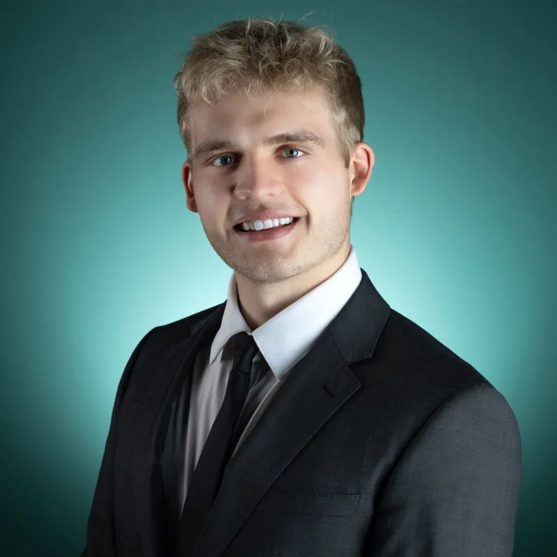 Young man with blond curly hair smiling in a dark suit and tie against a teal background