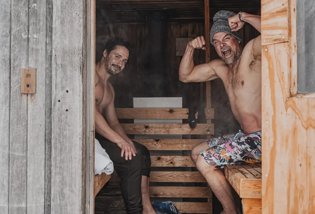 Two shirtless men sit inside a steam sauna, one flexing and shouting while wearing a knit beanie