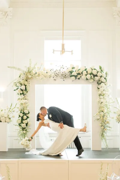Groom dipping bride for a kiss beneath a white floral arch at a wedding ceremony