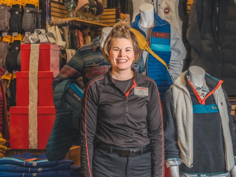 Smiling Scheels employee in a black uniform stands among outdoor apparel displays and mannequins