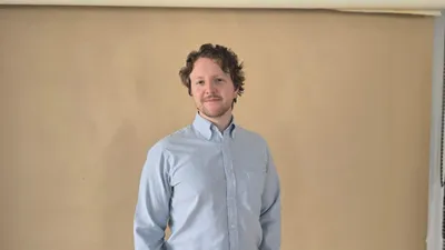 Smiling man with curly hair wearing a light blue button-up shirt against a beige wall