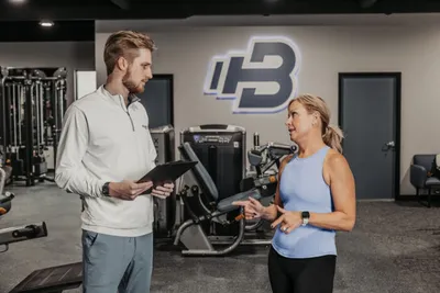 A trainer and a female client talk inside a gym with workout equipment