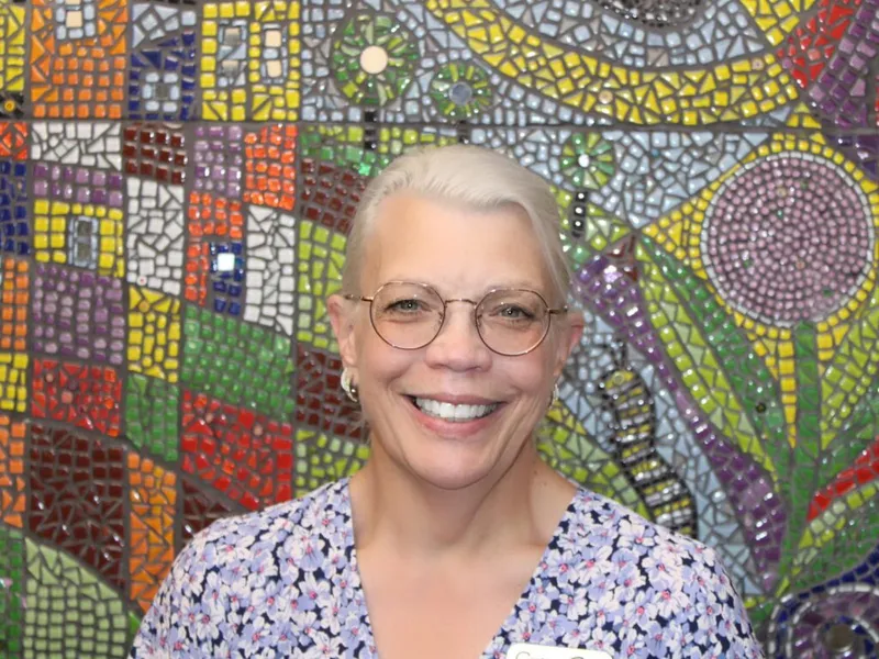 Smiling woman with white hair and round glasses in front of a colorful mosaic wall