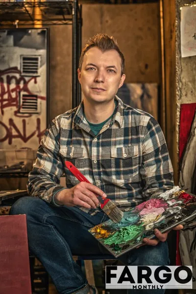 Man in plaid shirt holding a paintbrush and paint-covered palette in a graffiti-walled studio