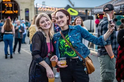 Two smiling women pose with drinks at an outdoor festival