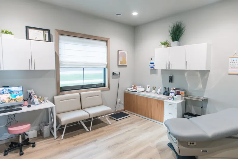 Bright medical exam room with beige chairs, exam table, white cabinets, and an eye chart