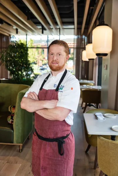 Red-bearded chef in white coat and burgundy apron stands arms crossed in a softly lit restaurant