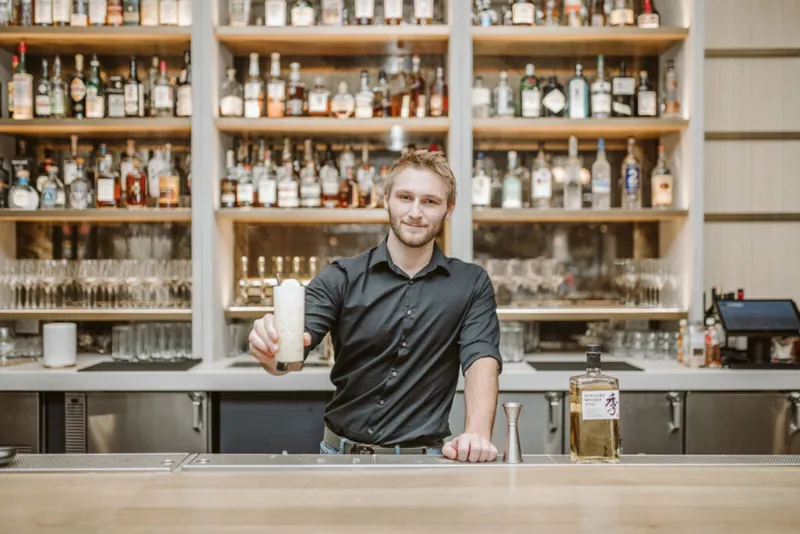 Smiling bartender in black shirt holds a frothy cocktail shaker behind a well-stocked bar