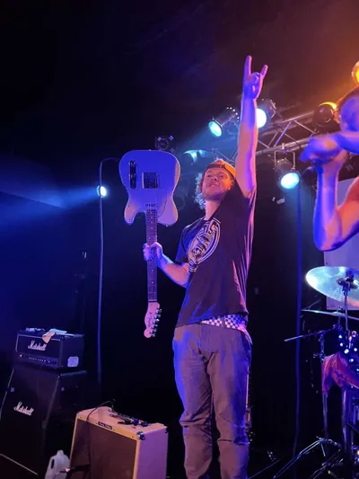 Guitarist raises devil horns onstage under blue stage lights, holding electric guitar at his side