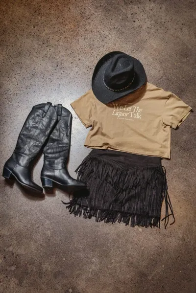 Flat lay of black cowboy boots, fringe mini skirt, tan graphic tee, and black felt hat