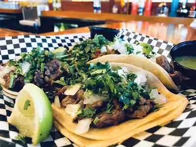 Three street tacos topped with cilantro and diced onion on a checkered wrapper with a lime wedge