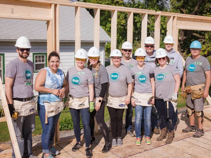Group of volunteers in white hard hats and matching T-shirts posing at a home construction site