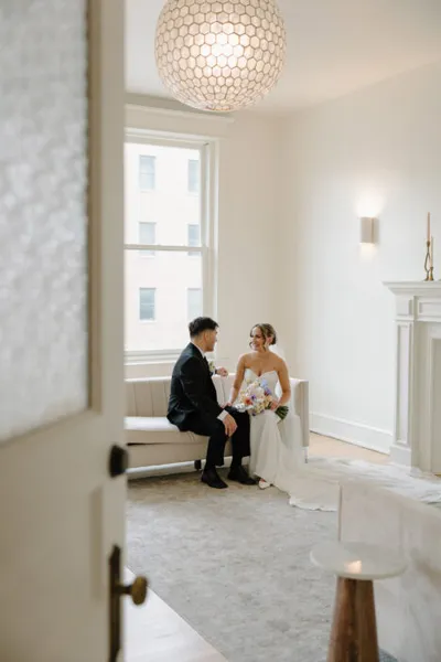 Bride in strapless gown holding bouquet chats with groom in dark suit by a bright window