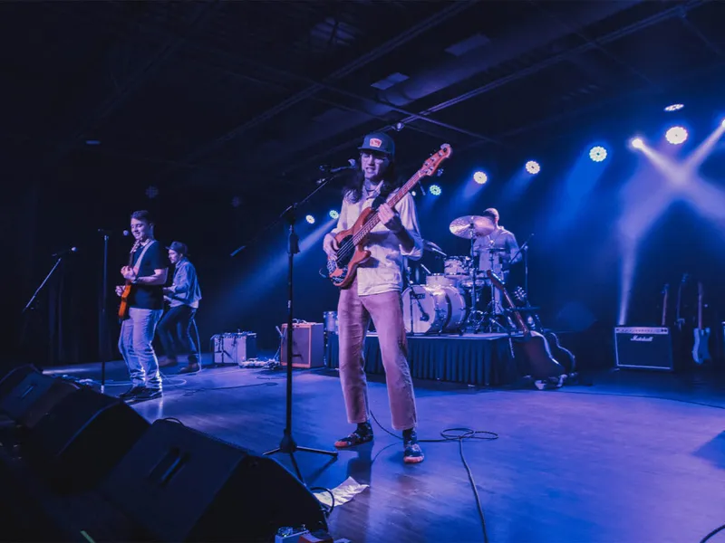 Band performs on a blue-lit stage, bassist in cap center with guitarist and drummer behind