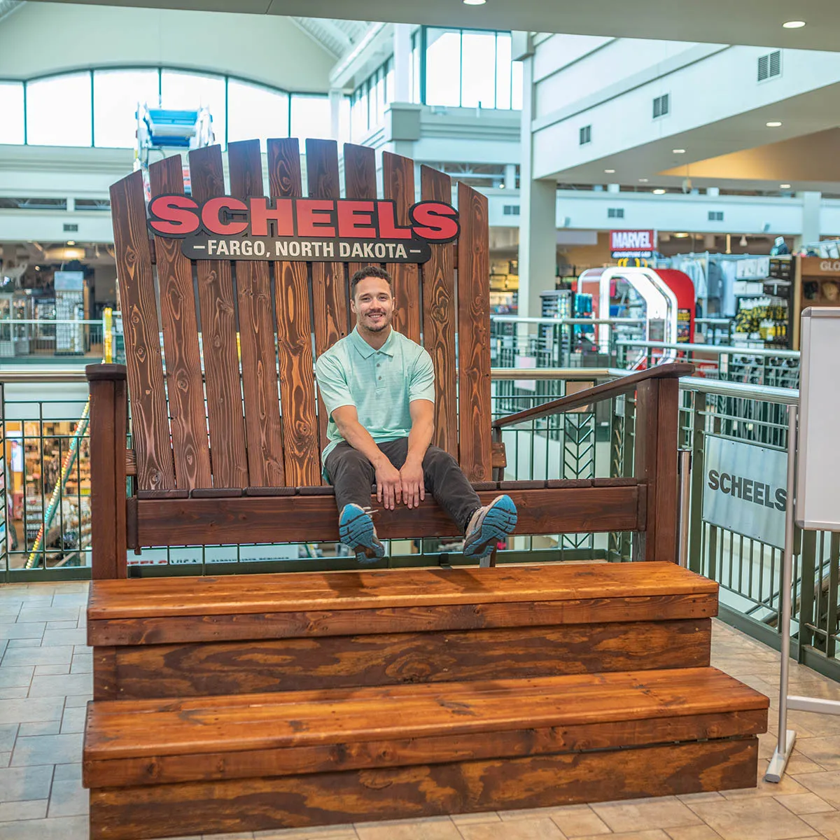 Man sitting in an oversized Adirondack chair branded Scheels Fargo, North Dakota, inside a retail store