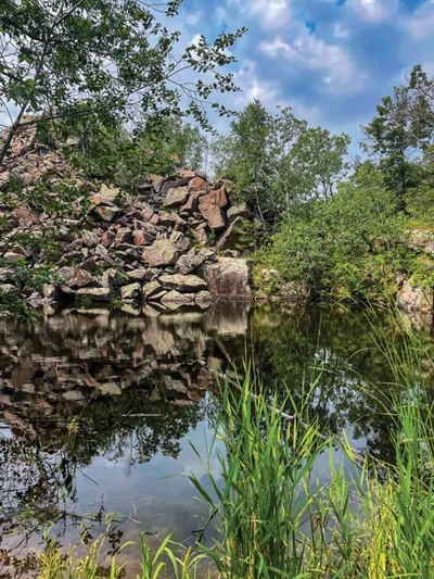 Rocky quarry wall reflected in still water surrounded by lush green trees