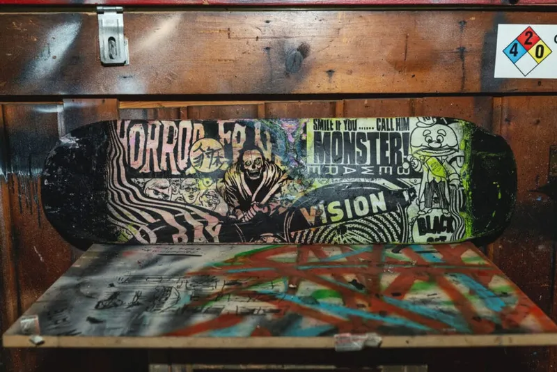 Decorated skateboard deck covered in horror-themed stickers and graffiti art resting on a wooden shelf