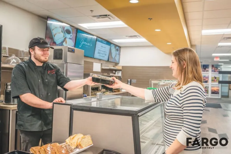 Smiling café worker in black chef uniform hands a packaged item to a woman across a deli counter