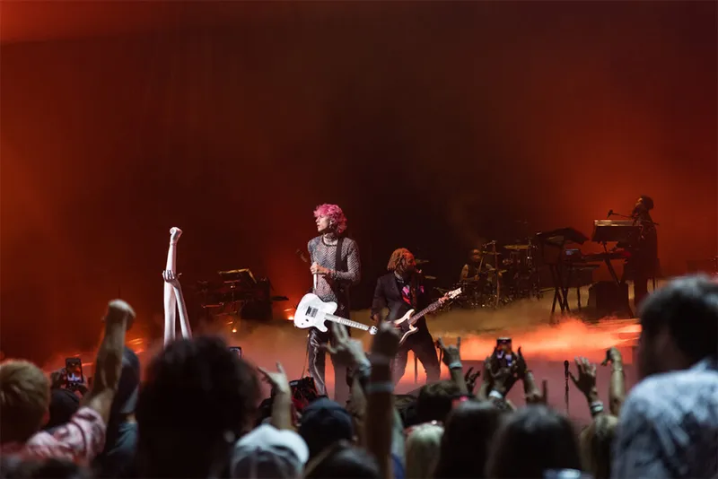 Pink-haired performer with white guitar on a fog-filled red-lit stage before a crowd