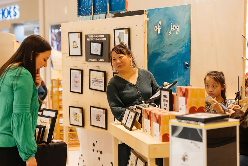 Smiling vendor at a craft fair booth displaying framed prints, greeting a browsing customer