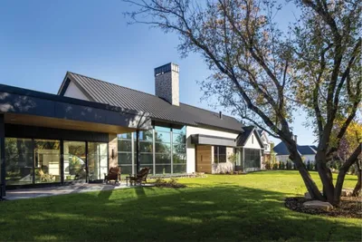 Modern farmhouse with black metal roof, glass walls, and lush green lawn framed by mature trees