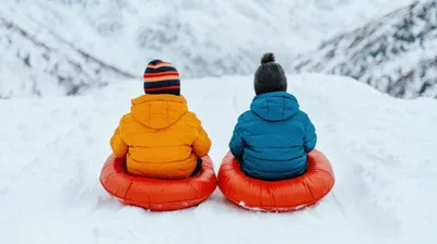 Two children in colorful winter jackets sitting on red snow tubes at the top of a snowy hill