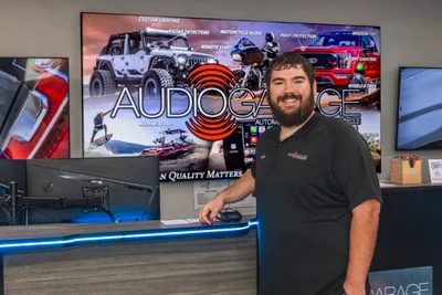 Smiling bearded man stands at a blue-lit counter in front of an Audio Garage branded display screen