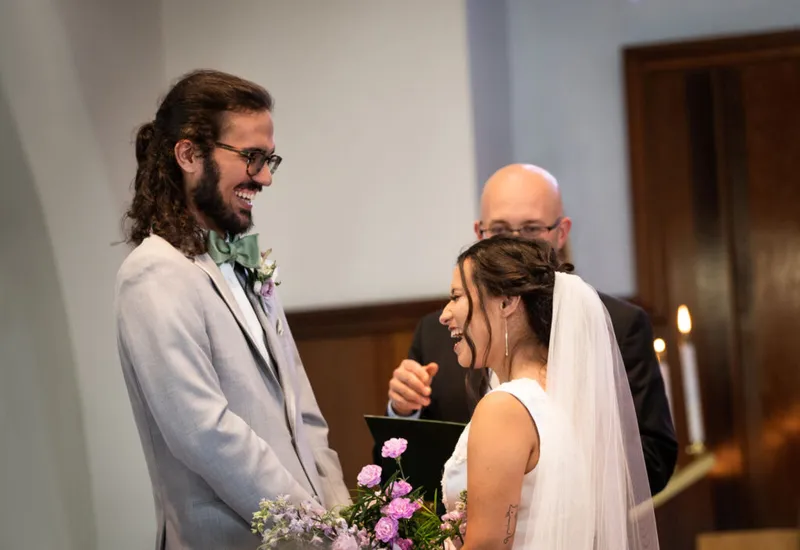 Laughing bride in white veil and groom in gray suit with green bow tie face each other at the altar