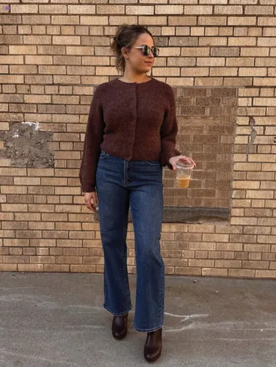 Woman in brown cardigan and wide-leg jeans holding an iced drink against a brick wall