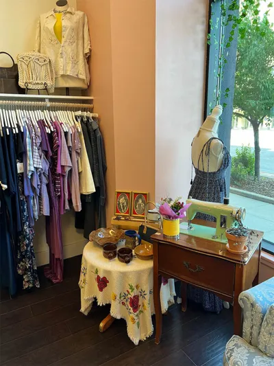 Vintage boutique interior with clothing rack, floral-draped side table, and dress form beside an antique sewing machine