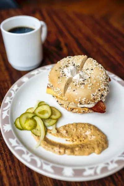 Everything bagel sandwich with bacon and cream cheese, served with pickles, dipping sauce, and black coffee