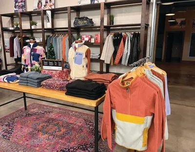 Clothing store display with folded garments on a wood table and colorful jackets on racks