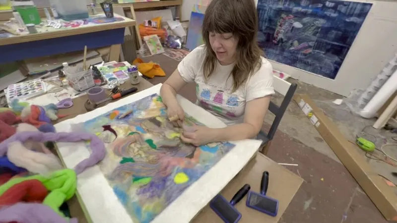 Woman in a white t-shirt works on a colorful mixed-media artwork in a cluttered art studio