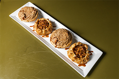 Four mini pies and rustic tarts on a white rectangular plate atop an olive green surface