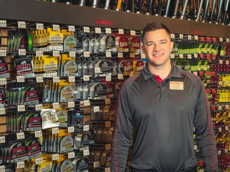 Scheels employee in branded polo smiling in front of a fishing line display wall