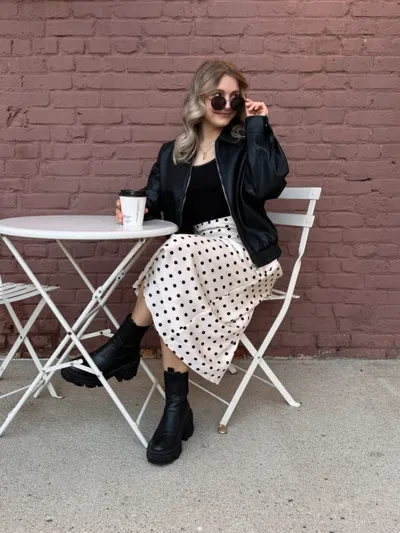 Woman in black leather jacket and polka-dot midi skirt seated at white bistro table against red brick wall