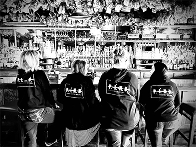 Four people in matching dark hoodies seated with backs to camera at a dimly lit bar