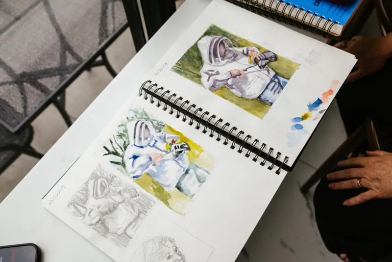 Open spiral sketchbook showing watercolor paintings and a pencil sketch of beekeepers