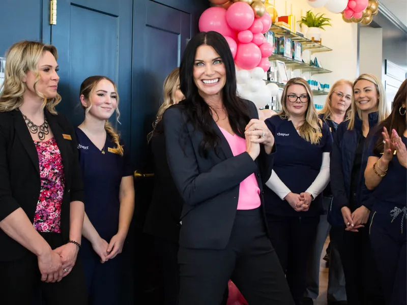 Smiling woman in black blazer and pink top stands before staff in navy scrubs, pink balloons behind
