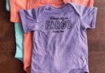 Purple Fargo graphic T-shirt laid flat on a wooden surface beside colorful shirts