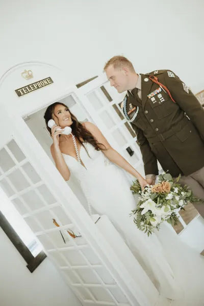 Bride in white gown on telephone booth phone, groom in military dress uniform holding bouquet