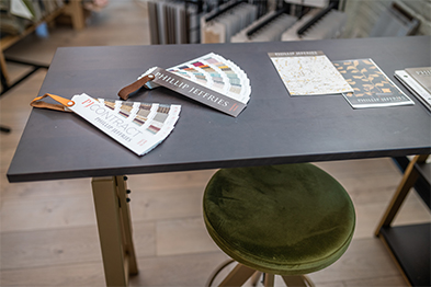 Fabric swatches and design catalogs spread on a dark worktable beside a green velvet stool